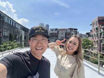 A selfie of a man wearing a 'Vietnam Veteran' cap and a woman smiling and gesturing with her hand, taken on a rooftop or elevated walkway in an urban area.