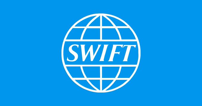 The image features a blue flag with white text that reads "SWIFT". The flag has a globe design at its center, which is surrounded by a border of gray lines. The background color of the flag is blue, providing a contrast to the white text on the flag. The image also includes some text in the bottom right corner that says "THIS IS BIG: SWIFT just completed ISO 20022 testing with Hedera, with rollout phases continuing through 2026." This information suggests that the flag might be related to an organization or project involving international standards and regulations.