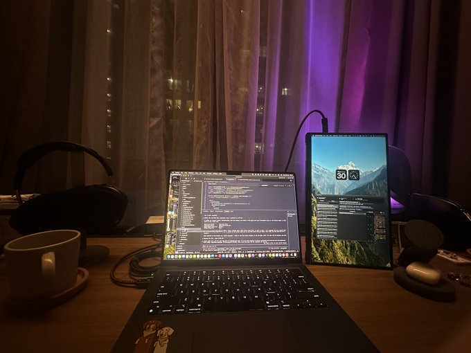 A dimly lit workstation setup featuring a laptop displaying code next to a tablet with a mountain wallpaper, accompanied by headphones, a coffee cup, and a microphone on a wooden desk.
