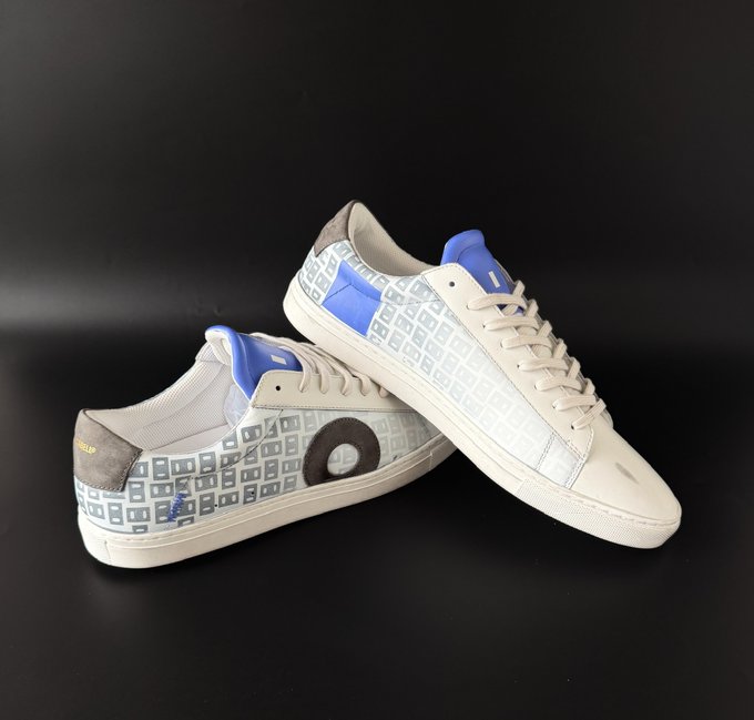 A pair of low-top sneakers in beige/cream and silver with blue accents on the heel tabs, featuring a patterned design and a circular logo on the side, photographed against a black backdrop.