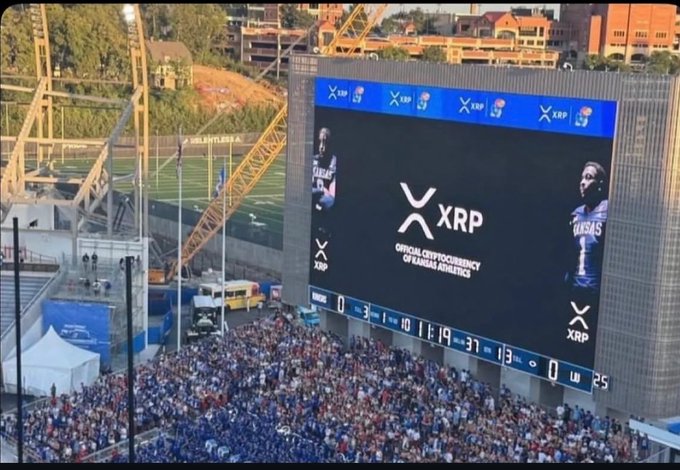 A massive stadium jumbotron displays an "XRP" logo declaring it the "Official Cryptocurrency of Kansas Athletics," flanked by two player graphics, while fans pack the stands below during a game showing a score of 0-37 in the first quarter. The image visually captures the XRP community's growth milestone referenced in the post, showcasing real-world brand adoption at a major college sports venue.