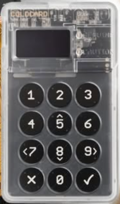 A transparent Coldcard hardware wallet displaying its numeric keypad with 0-9, special characters, and confirmation/cancel buttons.