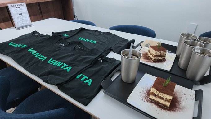A white table set with several dark t-shirts printed with 'VANTA' in teal, alongside two slices of tiramisu on white plates and metal cups with straws, with a menu stand visible in the background.