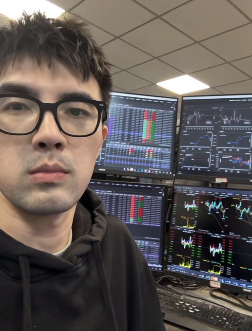 A young man with glasses takes a selfie in front of a multi-monitor trading setup displaying live cryptocurrency charts, candlestick patterns, and order book data—matching the post's claim about building a high-frequency trading bot that earned $208,452 on Polymarket in two and a half months.