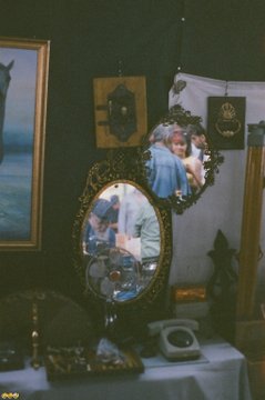 A display of framed mirrors and photos, with reflections showing visitors in a gallery or antique shop setting.