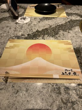 The image shows a dining table with two plates, one of which has an orange sun design on it. There are also chopsticks placed next to the plate with the sun design. The background appears to be a gray countertop or marble surface.