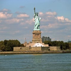 The Statue of Liberty stands prominently on Liberty Island, overlooking New York Harbor under a partly cloudy sky. The image symbolizes New York, the city considering extending its moratorium on Bitcoin mining to include AI data centers and other crypto operations.
