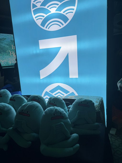 A row of plush toys with sad, downturned expressions sits in front of an illuminated blue banner featuring wave motifs and a large "ㅋ" character. The display, promoting "Hyperliquid," contrasts the cheerful branding with the somber faces of the stuffed figures, evoking the emotional weight often felt in volatile crypto and finance markets.