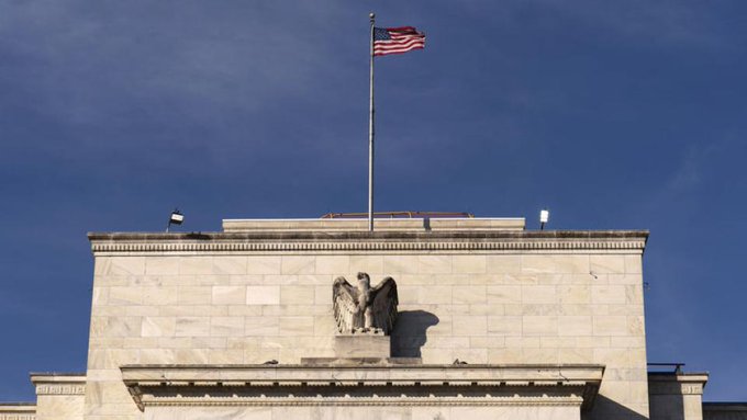 A stone building with an eagle sculpture on its facade is shown against a blue sky, with an American flag flying at half-mast on top.