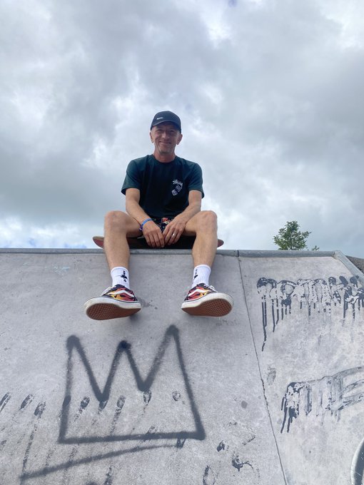 A man wearing a black cap, dark green t-shirt, white socks, and sneakers sits on the edge of a concrete skatepark ramp above graffiti, under a cloudy sky.