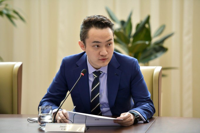 Justin Sun, the founder of TRON, wearing a blue suit and striped tie, sits at a conference table with a microphone and a glass of water, with a name placard partially reading 'JUSTIN CAI'.