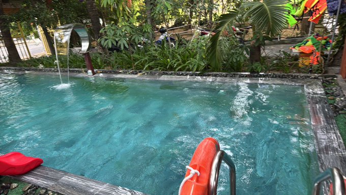 A clear turquoise swimming pool with a decorative water feature, surrounded by lush tropical greenery, with a red life preserver ring and red towel resting at the edge. The image conveys a relaxed, leisurely Sunday atmosphere, fitting the post's casual tone about chilling out rather than focusing on cryptocurrency activity.