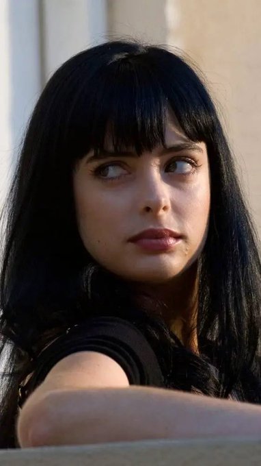 A portrait of a young woman with long dark hair and bangs, looking to the side with a subtle smile, wearing a black top.