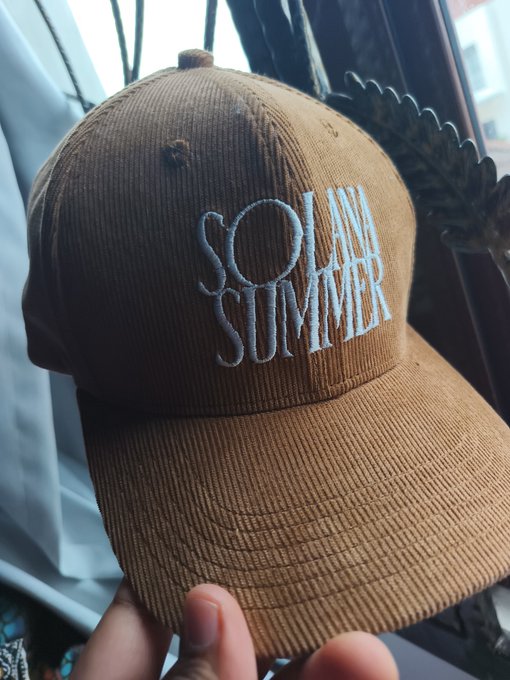 A hand holds up a brown corduroy baseball cap with white embroidered text reading 'SO AM SUMMER' against a background of curtains and a window.
