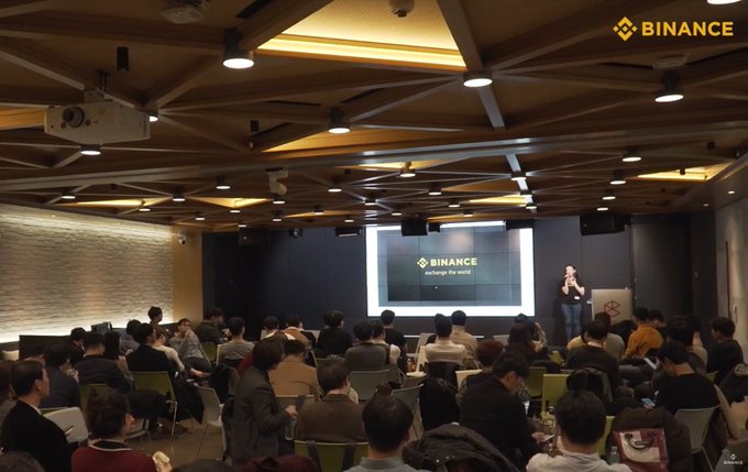 A presenter addresses a packed audience at the BNBChain Builders gathering during Korea Blockchain Week 2026 in Seoul, with the Binance logo prominently displayed on a large projection screen showing "exchange the world." The scene captures the modern, large-scale evolution of Binance's presence in Korea, contrasting with the intimate 2018 meetup referenced in the post.