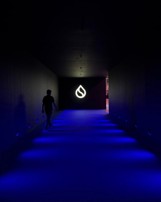 A silhouetted figure walks down a dark corridor illuminated by blue lights on the floor, leading toward a bright white liquid drop logo on the back wall.