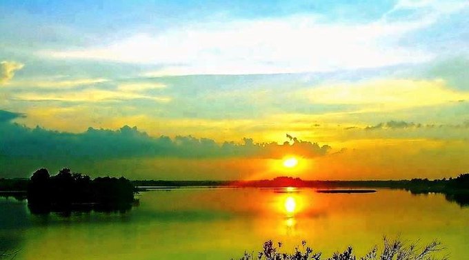 A scenic landscape photograph of a vibrant sunset over a calm lake, with the sun's golden reflection on the water and a dramatic sky of yellow and blue hues.