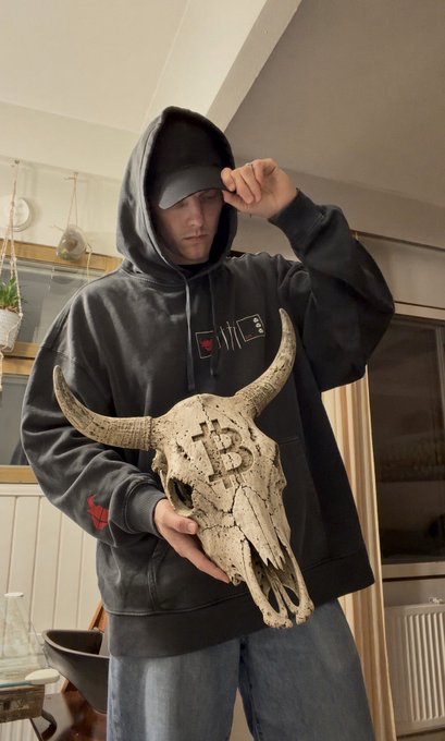A person wearing a dark gray hoodie and cap holds a weathered animal skull with long horns that has a Bitcoin symbol carved into its forehead.