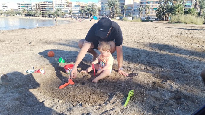 Father and young child playing in the sand at a beach with colorful toys including a red shovel, with water and apartment buildings visible in the background.