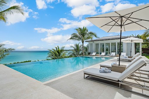 Luxurious tropical resort scene featuring an infinity pool overlooking turquoise ocean waters, with palm trees, white lounge chairs, and umbrellas under a bright blue sky.