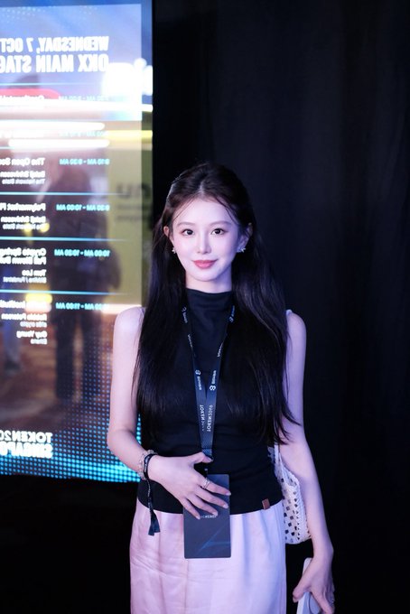 A young woman with long dark hair, wearing a black sleeveless top and white skirt with a lanyard, poses in front of a schedule board listing events like 'OHM MAIN STAGE' and 'MIDNIGHT CO.', holding a smartphone.
