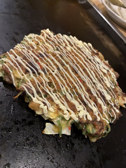 A large rectangular Japanese okonomiyaki or savory pancake topped with mayonnaise drizzle and bonito flakes on a black griddle.