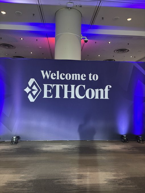 The image shows a stage with a large blue sign that reads "Welcome to EthConf", indicating an event or gathering related to ethical discussions, conferences, or workshops. The stage appears to be set up for presentations or performances, as there are two speakers positioned on either side of the stage and a microphone is visible in front of them.