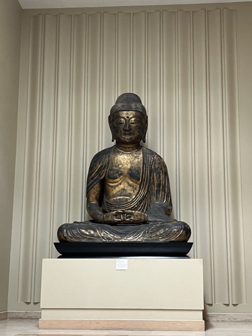 A large bronze Buddha statue seated in a meditation pose on a pedestal against a backdrop of beige curtains.