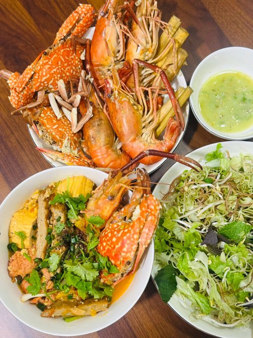 A spread of seafood including blue swimmer crabs, large prawns, a bowl of green soup, a salad with bean sprouts and herbs, and a bowl of fish.