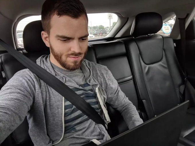 The image shows a man sitting inside a car with his laptop open in front of him. The man has a beard, and he appears to be focused on something on his computer screen. There are no other people or objects visible in the scene. The photo is taken from the back seat of the car, providing an interesting perspective on the man's activity while traveling.