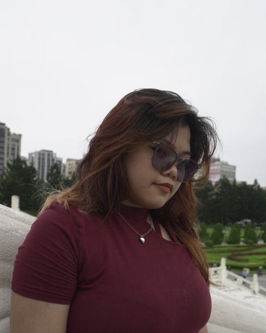 A selfie-style photo of a woman with wavy brown hair wearing purple-tinted sunglasses and a maroon shirt, with buildings and greenery visible in the background.