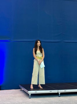 A young woman stands barefoot on a small stage, holding a light blue cloth, delivering a presentation against a blue backdrop at the @ethbelgrade event. Her poised stance and the setup suggest she is introducing the first day of talks covering Ethereum's state, the future of Solidity, and Glamsterdam.