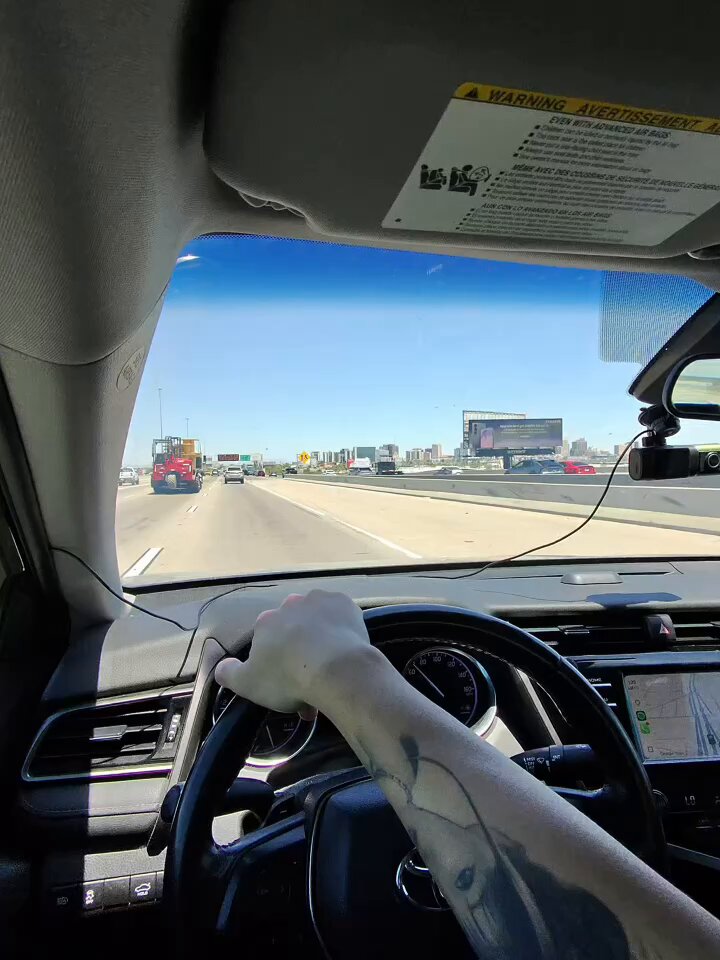 The image shows a person driving a car with their hand on the steering wheel, likely taking part in an onboarding video or promotional activity for a company named "Core". The driver's tattoo can be seen on their arm as they navigate through traffic.