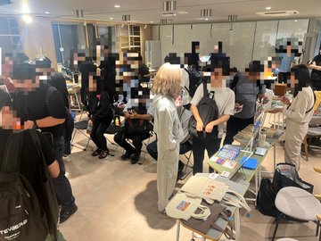 Crowded indoor scene with people whose faces are blurred, one person with light hair wearing a white top in the center, with tables displaying merchandise or magazines.