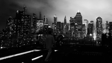The image shows a city skyline at night, featuring tall buildings illuminated by streetlights. A person can be seen walking across the scene, adding life to the otherwise dark surroundings. The photo appears to have been taken during the day, as indicated by the presence of daylight and the fact that it is nighttime in the background.