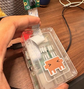 A person's hands interact with a Raspberry Pi single-board computer housed in a clear plastic case adorned with an orange cartoon cat sticker, featuring a small cooling fan and attached camera module.