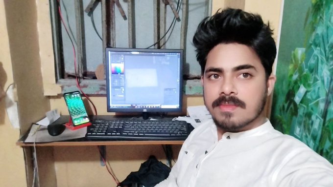 A young man with styled hair and a mustache sitting at a desk with a computer monitor, keyboard, and a smartphone displaying a green screen.