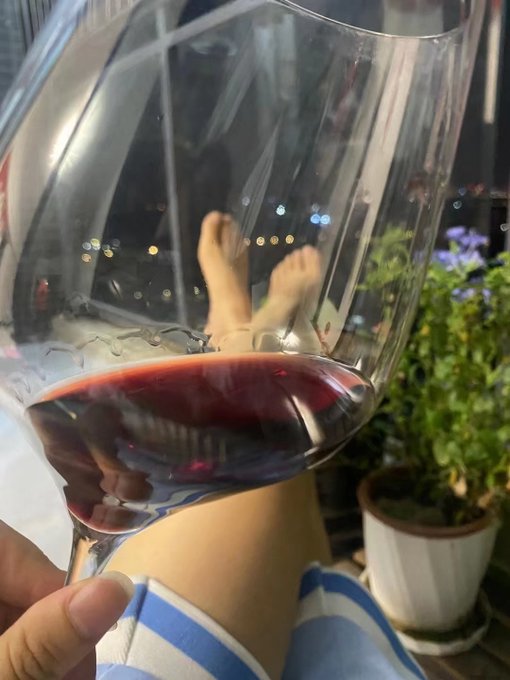 A hand holds a wine glass of red wine, with the photo taken through the glass showing a reflection of someone's feet crossed at the ankle against a night cityscape backdrop, alongside a potted plant. The post references Bitcoin price predictions ("大饼" meaning "big pie," a nickname for BTC) and weekend market consolidation around the 58,000 level, pairing the relaxing wine moment with a casual commentary on crypto market sentiment.