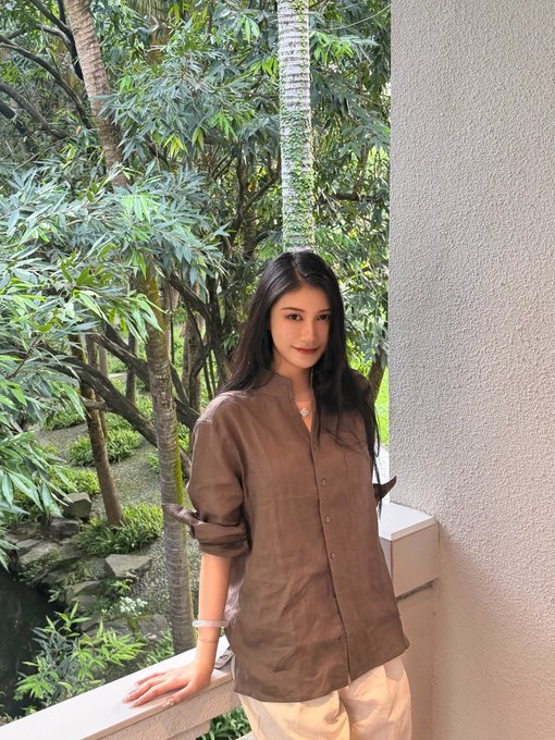 A woman with long dark hair wearing a brown button-up shirt and light pants, leaning against a ledge with lush green trees behind her.