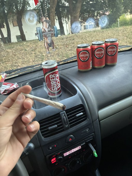 A hand holds a lit joint inside a car, with a 'Super Bock' beer can in a cupholder and three 'Zambujeira' beer cans on the dashboard.