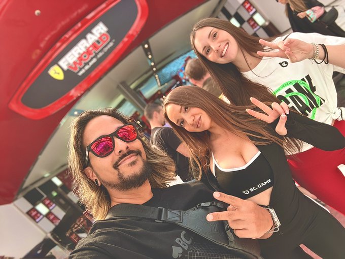 A man in red mirrored sunglasses takes a selfie with two smiling women at the Ferrari World booth, all flashing peace signs in support of the BC.GAME family as indicated by the logo on the woman's top and the "BC.GAME" text visible on the Ferrari World signage.