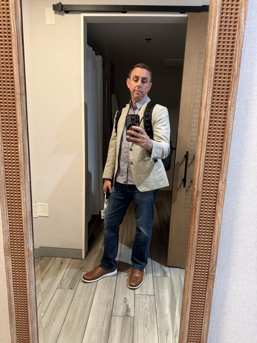 A man takes a mirror selfie wearing a beige blazer, patterned shirt, jeans, brown shoes, and a backpack, standing on wood-look tile flooring.