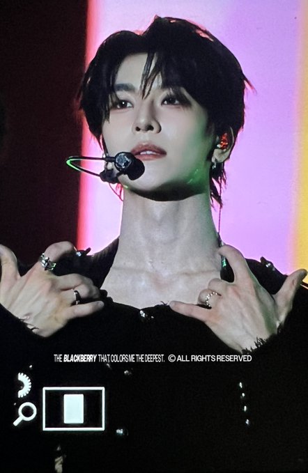 A young male performer with dark hair and an earpiece microphone poses dramatically with both hands near his collar, wearing a black outfit and multiple rings. The image is watermarked with "THE BLACKBERRY THAT COLOR ME THE DEEPEST © ALL RIGHTS RESERVED" and shows an on-screen interface with a magnification icon. The accompanying post tags suggest this is a fan photo from a concert or performance event, with the user asking about the situation likely referring to the performer's striking pose or expression captured in the moment.
