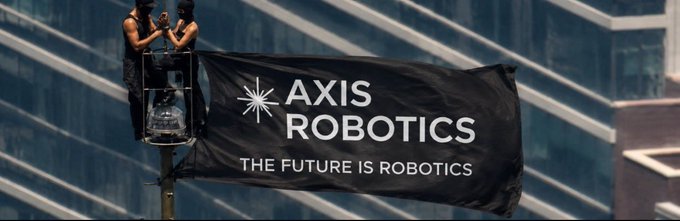Two workers on a lift hang a large black banner reading "AXIS ROBOTICS - THE FUTURE IS ROBOTICS" against the backdrop of a modern glass building, visually marking the company's branding milestone mentioned in the post about Axis Robotics expanding its Physical AI initiatives.