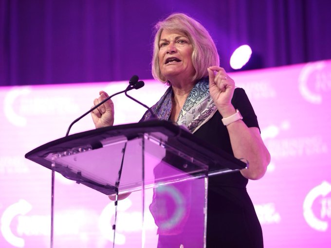 Senator Cynthia Lummis stands at a clear podium gesturing with both hands as she speaks at a podium backed by purple curtains and pink "FIRST" branded signage, illustrating her pointed remarks attributing potential failure of the CLARITY Act to Democrats after months of negotiation.