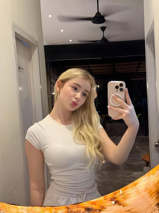 A mirror selfie of a young blonde woman wearing a white t-shirt and light shorts, posing with puckered lips while holding a phone with a ring holder, in a modern interior with ceiling fans visible behind her.