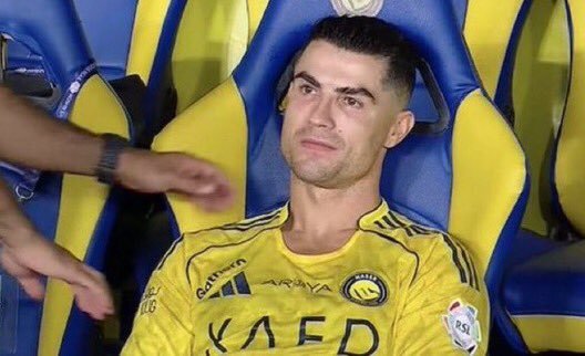 A photo of soccer player Cristiano Ronaldo wearing a yellow Al Nassr jersey with 'KAED' visible, sitting on the bench with a focused expression during a match.