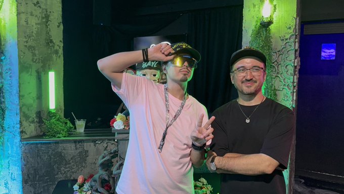 Two men pose together indoors, one wearing a pink shirt, sunglasses, and a cap, the other in a black t-shirt and glasses, with neon lights in the background.