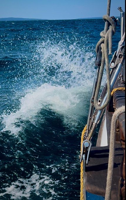 A view from the side of a boat, showing ocean waves crashing and churning against the hull near ropes and metal hardware.