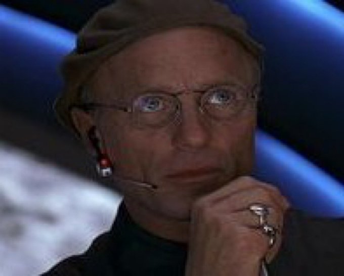 A close-up shot shows actor Christoph Waltz in a role, wearing glasses, a cap, and a headset, looking intently to the side.